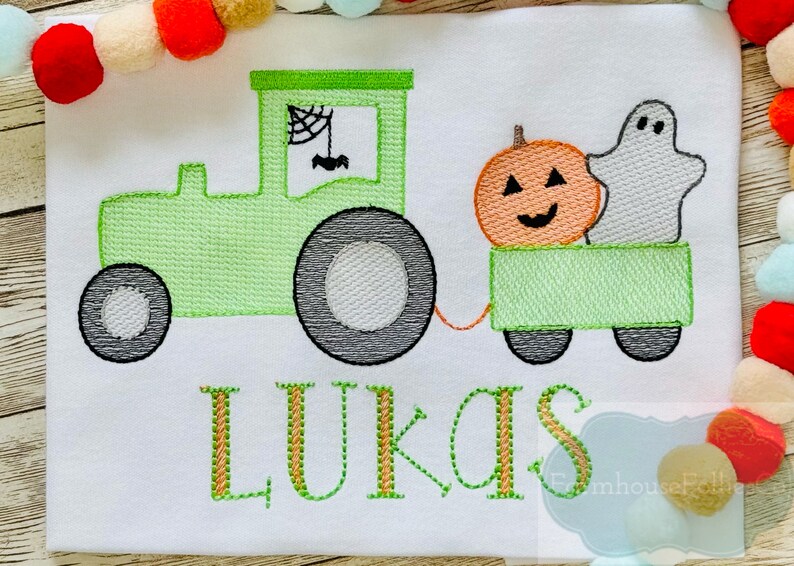 Op de afbeelding: Een groene tractor trekt een wagen met een pompoen en een spook. De wagen is groen en heeft zwarte wielen. De tractor heeft een spinnenweb in het raam. De naam "LUKAS" is in oranje en groen geborduurd aan de onderkant van de afbeelding.