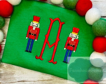 May include: Green Christmas shirt featuring two embroidered nutcracker figures and a large letter "M" in red thread. The shirt is surrounded by red, white and green pom-poms.