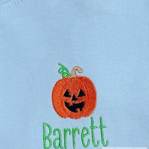 May include: Light blue shirt with an embroidered orange jack-o'-lantern and the name "Barrett" in green script. The pumpkin has a carved face and a green vine. The shirt is a light blue color.