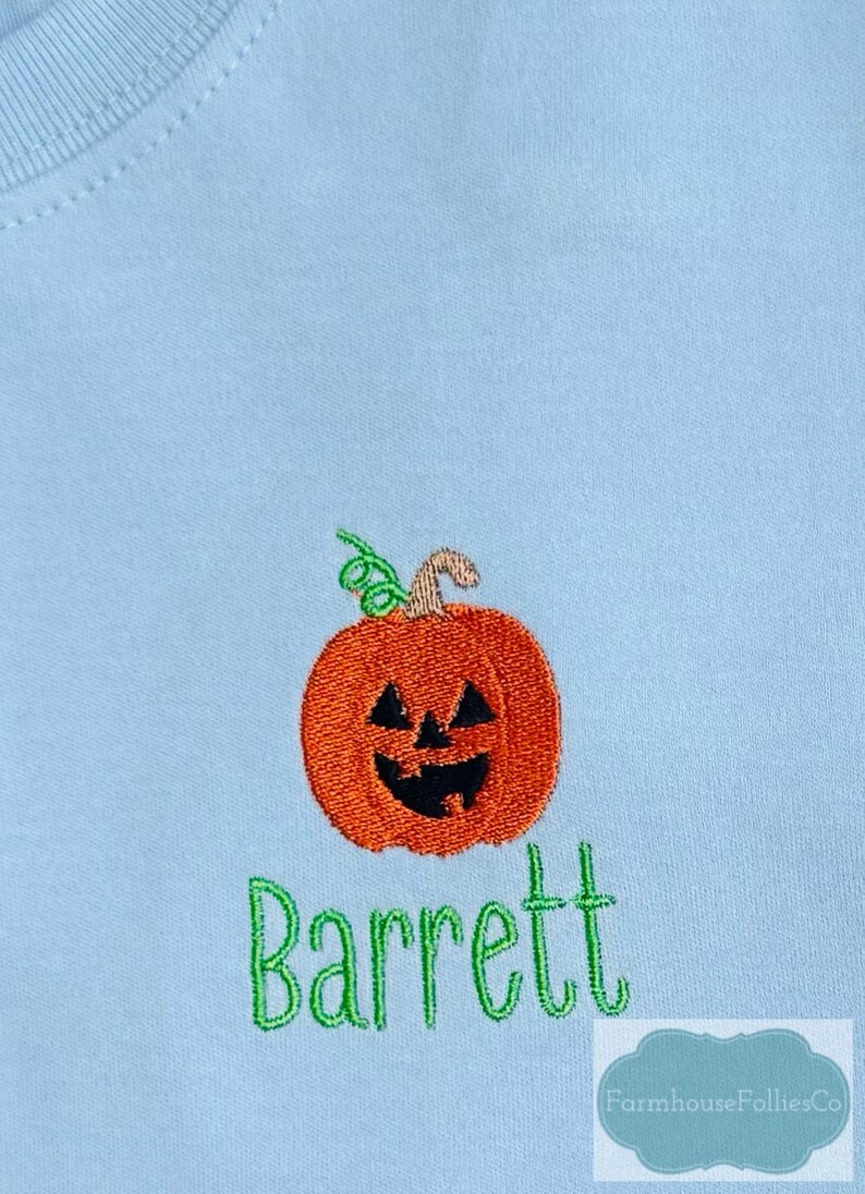 May include: Light blue shirt with an embroidered orange jack-o'-lantern and the name "Barrett" in green script. The pumpkin has a carved face and a green vine. The shirt is a light blue color.