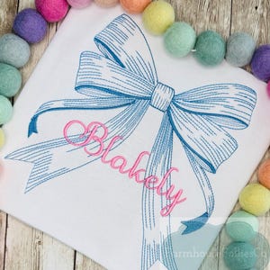 May include: White fabric with a large blue bow design and the name "Blakely" in pink script. The bow is detailed with fine lines. A garland of colorful felt balls surrounds the fabric.