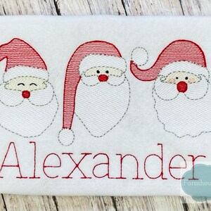 K&ouml;nnte beinhalten: Gestickte Darstellung von drei Weihnachtsmann-Gesichtern mit roten M&uuml;tzen und wei&szlig;en B&auml;rten. Der Name "Alexander" ist unter den Gesichtern in roter Farbe gestickt.
