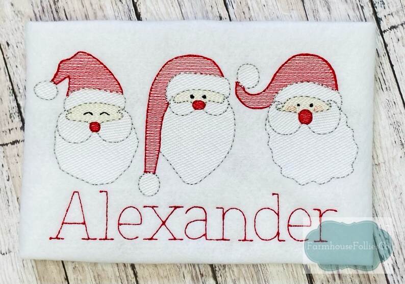 K&ouml;nnte beinhalten: Gestickte Darstellung von drei Weihnachtsmann-Gesichtern mit roten M&uuml;tzen und wei&szlig;en B&auml;rten. Der Name "Alexander" ist unter den Gesichtern in roter Farbe gestickt.