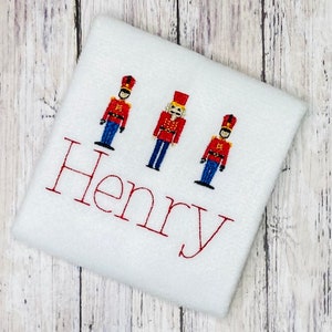 May include: White fabric with three embroidered toy soldier figures and the name "Henry" in red thread.