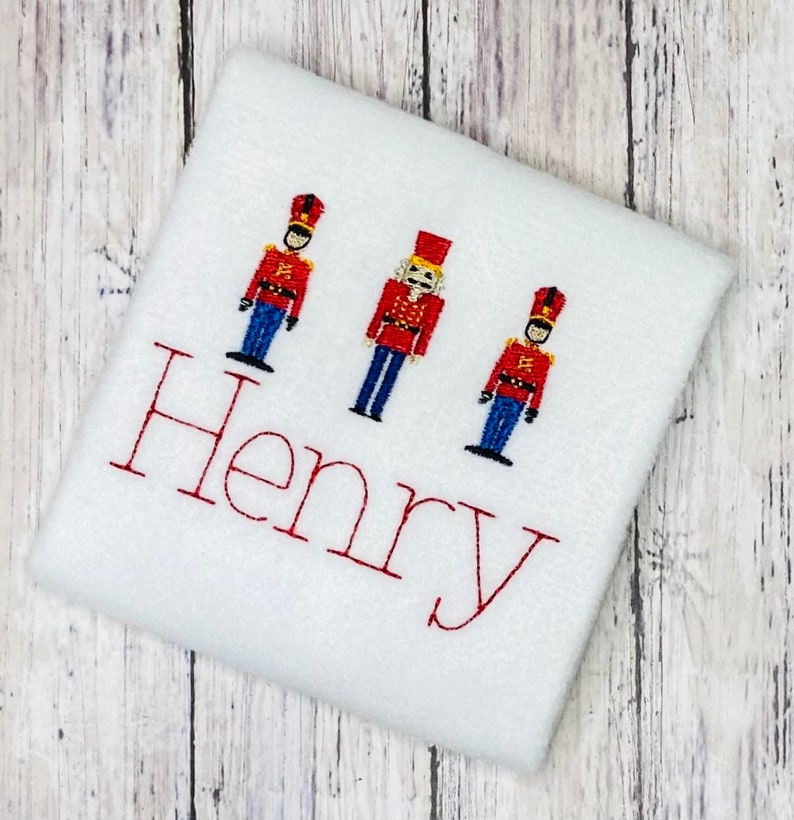 May include: White fabric with three embroidered toy soldier figures and the name "Henry" in red thread.
