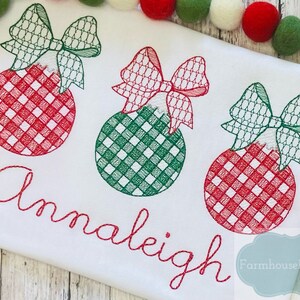 Pode incluir: Uma camiseta branca com enfeites de Natal bordados em vermelho e verde e o nome "Annaleigh" em bordado vermelho. O texto "Farmhouse Follies Co." tamb&eacute;m est&aacute; bordado em vermelho no canto inferior direito da camiseta.