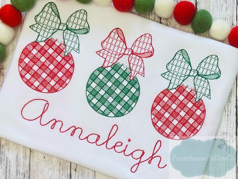 Pode incluir: Uma camiseta branca com enfeites de Natal bordados em vermelho e verde e o nome "Annaleigh" em bordado vermelho. O texto "Farmhouse Follies Co." tamb&eacute;m est&aacute; bordado em vermelho no canto inferior direito da camiseta.