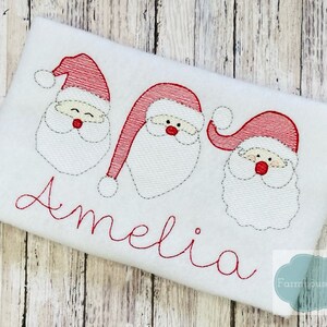 May include: White embroidered fabric with three red and white Santa faces and the name Amelia in red cursive lettering.