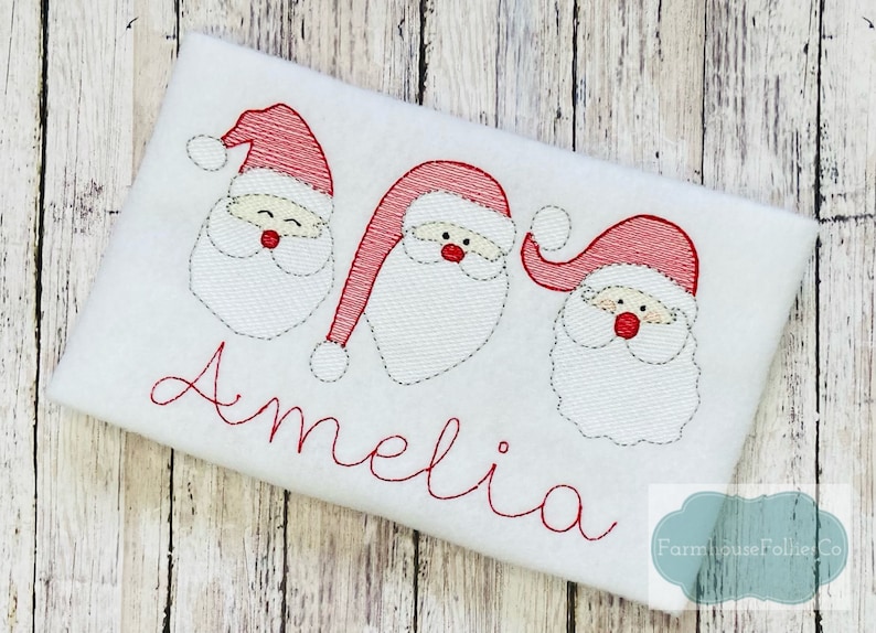 May include: White embroidered fabric with three red and white Santa faces and the name Amelia in red cursive lettering.