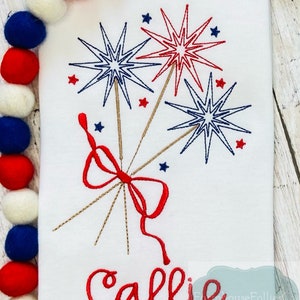 May include: A white t-shirt with a red, white, and blue embroidered design of fireworks and a bow. The name "Callie" is embroidered in red below the design.
