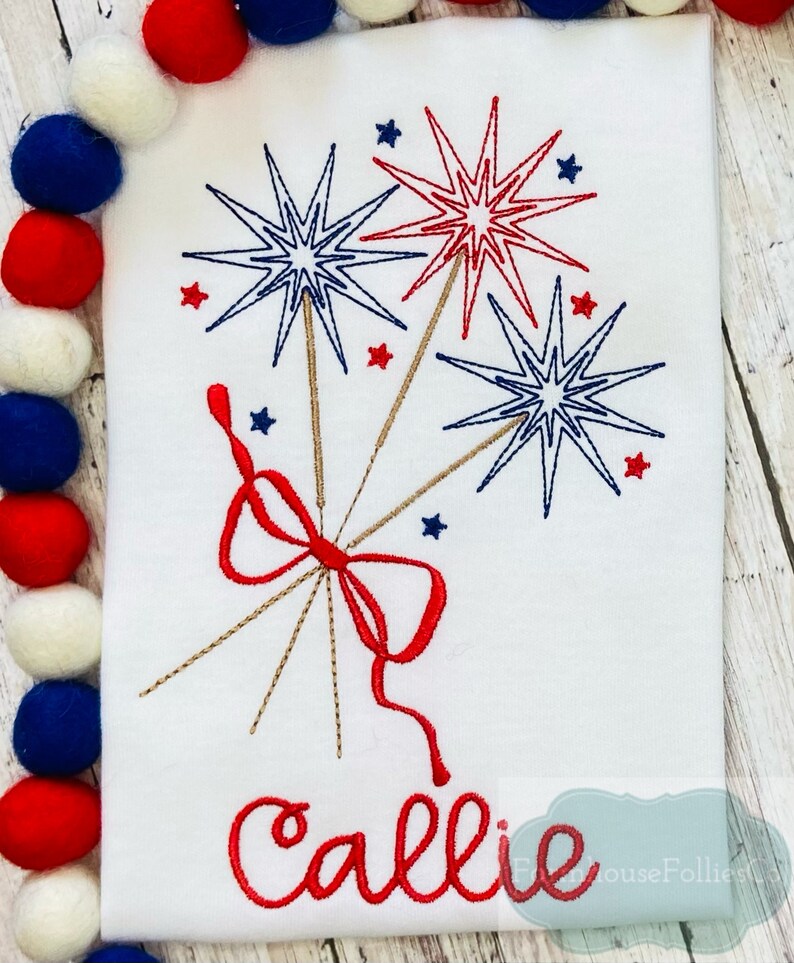 May include: A white t-shirt with a red, white, and blue embroidered design of fireworks and a bow. The name "Callie" is embroidered in red below the design.