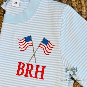 K&ouml;nnte beinhalten: Hellblau-wei&szlig; gestreiftes Kleinkind-T-Shirt mit gekreuzten, rot-wei&szlig;-blau gestickten amerikanischen Flaggen. Das Shirt hat die gestickten Buchstaben "BRH" in Rot. Gr&ouml;&szlig;e 2t.