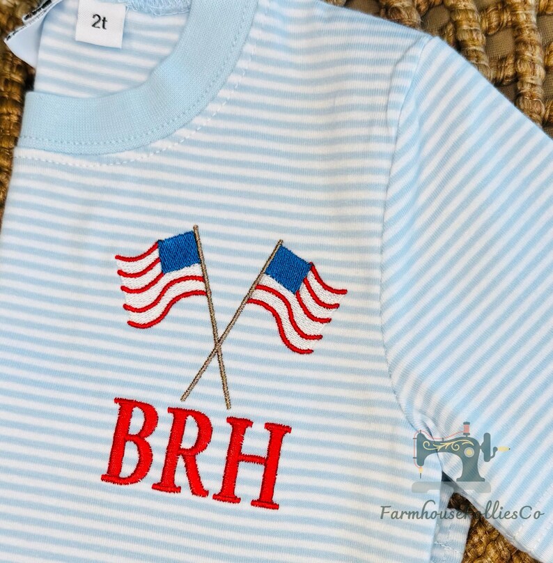 K&ouml;nnte beinhalten: Hellblau-wei&szlig; gestreiftes Kleinkind-T-Shirt mit gekreuzten, rot-wei&szlig;-blau gestickten amerikanischen Flaggen. Das Shirt hat die gestickten Buchstaben "BRH" in Rot. Gr&ouml;&szlig;e 2t.