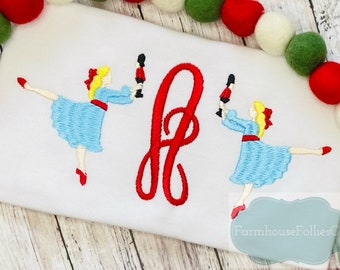 May include: Embroidered design of two ballerinas in blue dresses holding toy soldiers, with a red cursive letter "F" in the center. The design is on a white fabric background.