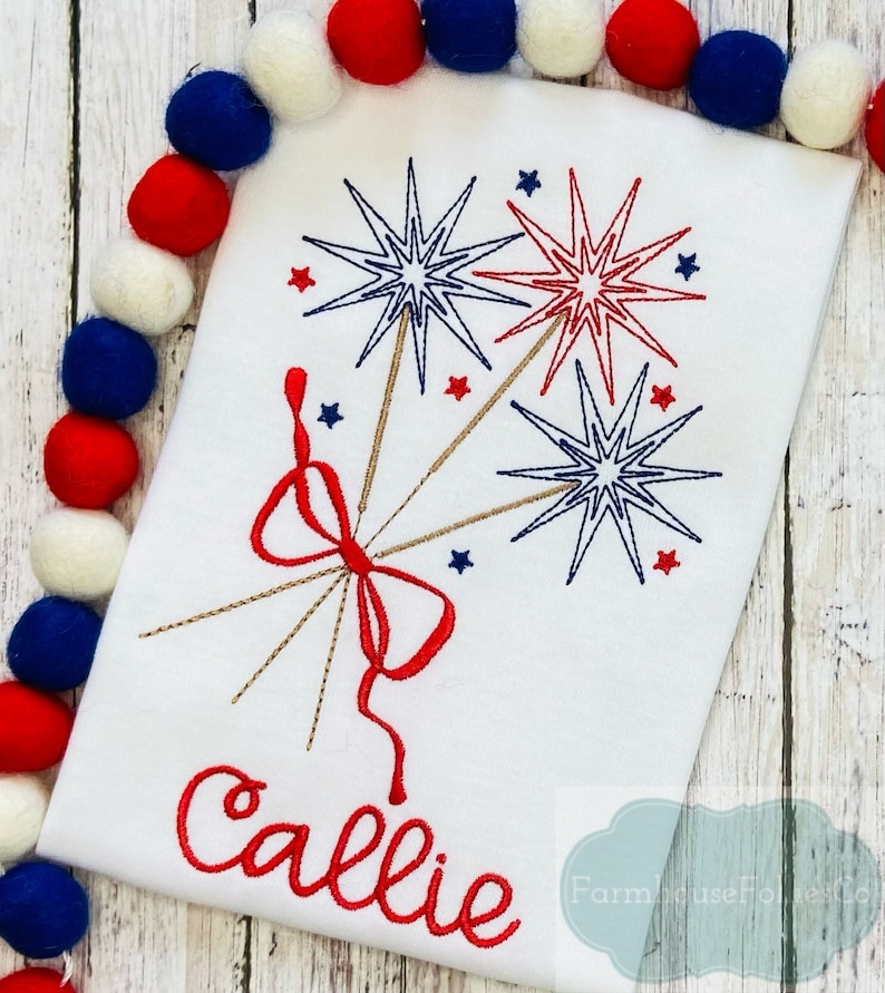 May include: A white t-shirt with a red, white, and blue embroidered design of fireworks and a bow. The name "Callie" is embroidered in red below the design.