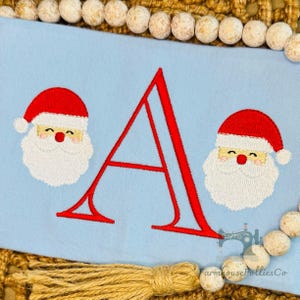Pode incluir: Um painel de tecido azul-claro com uma letra A grande bordada em vermelho, centralizada entre dois rostos de Papai Noel bordados. Cada Papai Noel tem um gorro vermelho, barba branca e bochechas rosadas. O painel é emoldurado por contas claras.