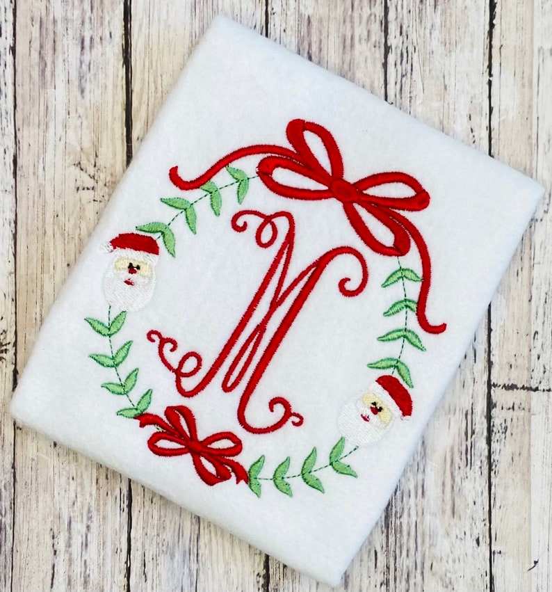 Puede incluir: Guirnalda de Navidad bordada con un lazo rojo, hojas verdes y dos caras de Pap&aacute; Noel que rodean la letra M.
