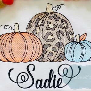 May include: Embroidered design featuring three pumpkins with a leopard print, orange, and blue color scheme. The name "Sadie" is embroidered below the pumpkins.