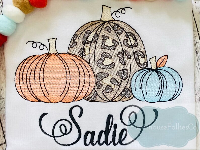 May include: Embroidered design featuring three pumpkins with a leopard print, orange, and blue color scheme. The name "Sadie" is embroidered below the pumpkins.