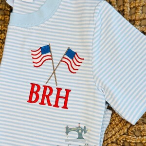 K&ouml;nnte beinhalten: Hellblau-wei&szlig; gestreiftes T-Shirt mit gestickten amerikanischen Flaggen und den Buchstaben "BRH" in Rot. Das Shirt hat einen hellblauen Kragen und kurze &Auml;rmel. Das Etikett lautet "2t".