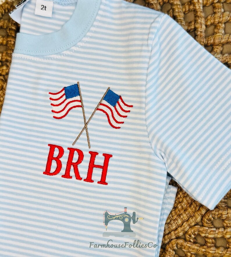 K&ouml;nnte beinhalten: Hellblau-wei&szlig; gestreiftes T-Shirt mit gestickten amerikanischen Flaggen und den Buchstaben "BRH" in Rot. Das Shirt hat einen hellblauen Kragen und kurze &Auml;rmel. Das Etikett lautet "2t".