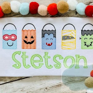 May include: A white t-shirt with an embroidered design of five Halloween treat bags with different character faces. The bags are in a row and the name "Stetson" is embroidered below them.