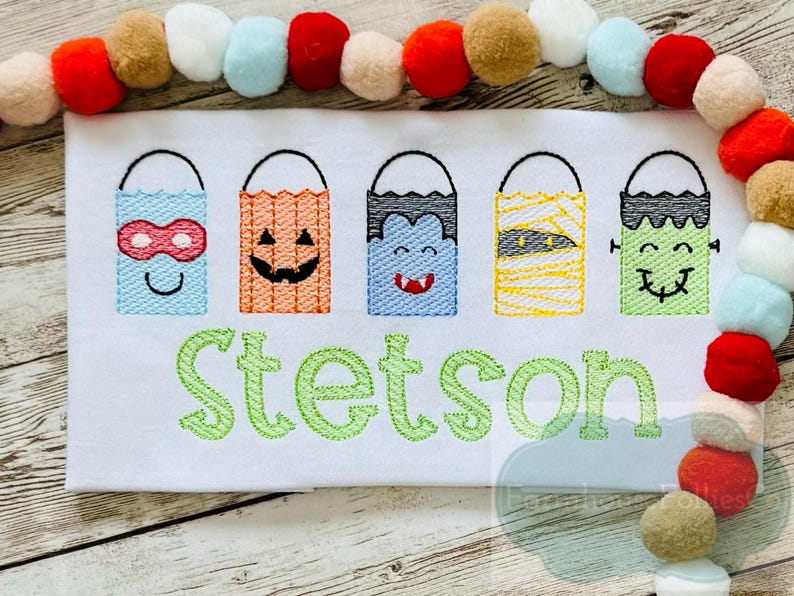 May include: A white t-shirt with an embroidered design of five Halloween treat bags with different character faces. The bags are in a row and the name "Stetson" is embroidered below them.