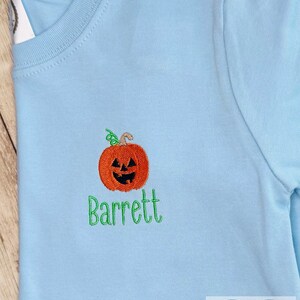 May include: Light blue long-sleeved shirt with an embroidered orange pumpkin and the name "Barrett" in green. The pumpkin has a carved face and a green stem. The shirt is a crew neck style.