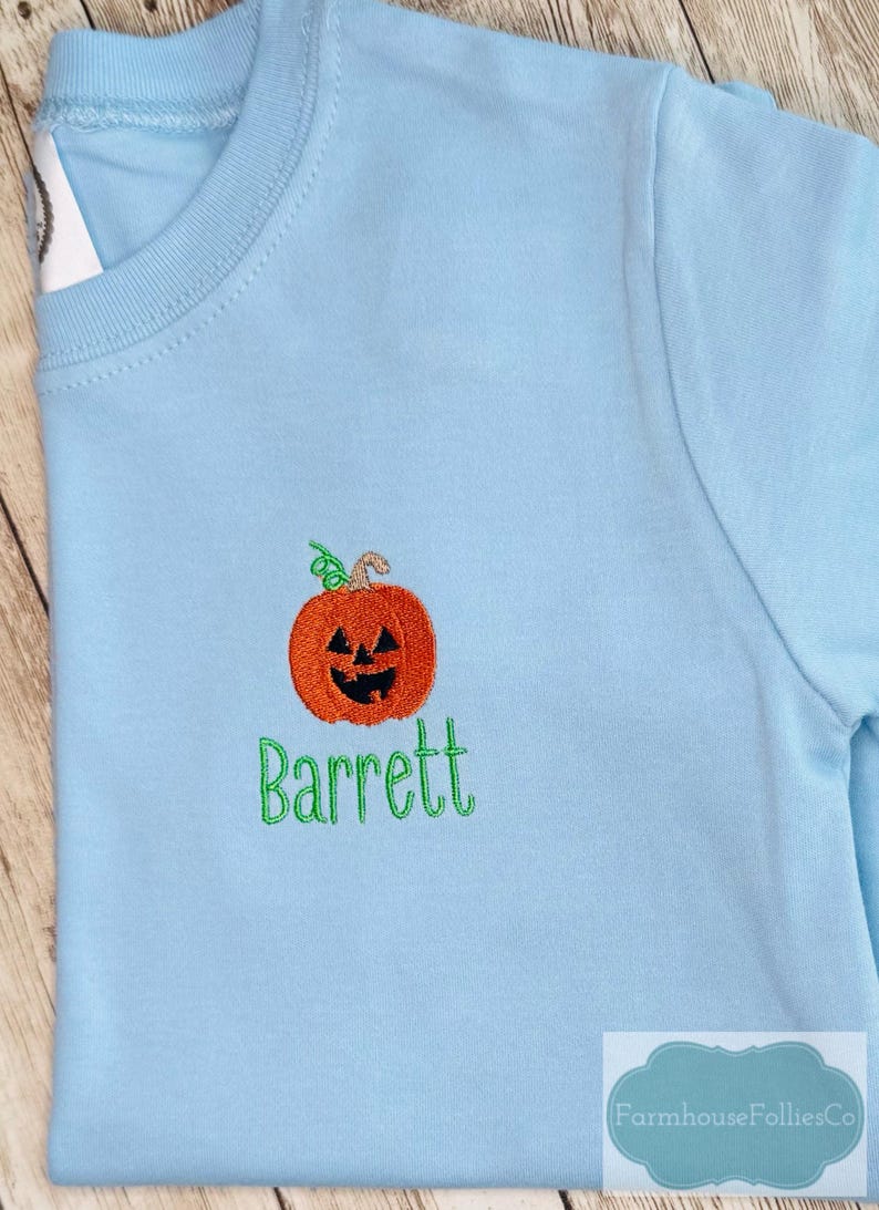 May include: Light blue long-sleeved shirt with an embroidered orange pumpkin and the name "Barrett" in green. The pumpkin has a carved face and a green stem. The shirt is a crew neck style.