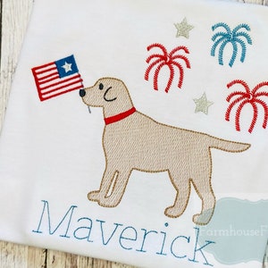 Op de afbeelding: Een wit T-shirt met een geborduurd beeld van een gele Labrador Retriever die een Amerikaanse vlag in zijn bek houdt. Rondom de hond zijn rode, witte en blauwe vuurwerkjes geborduurd. De naam "Maverick" is onder de hond geborduurd.