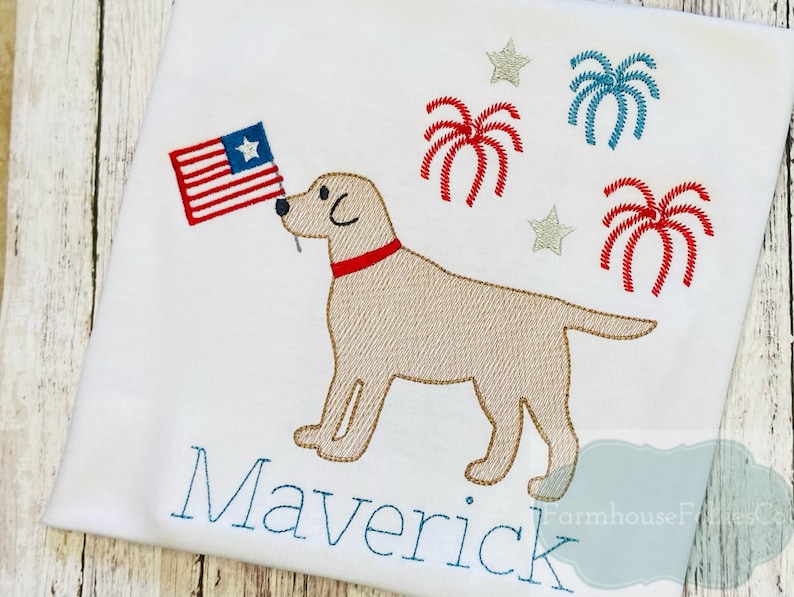 Op de afbeelding: Een wit T-shirt met een geborduurd beeld van een gele Labrador Retriever die een Amerikaanse vlag in zijn bek houdt. Rondom de hond zijn rode, witte en blauwe vuurwerkjes geborduurd. De naam "Maverick" is onder de hond geborduurd.