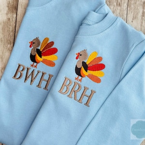 Pode incluir: Duas camisas de manga comprida azul claro com desenhos de peru bordados. Cada peru tem um corpo castanho, penas da cauda coloridas e uma barbela vermelha. As camisas têm as iniciais bordadas "BWH" e "BRH" em castanho.