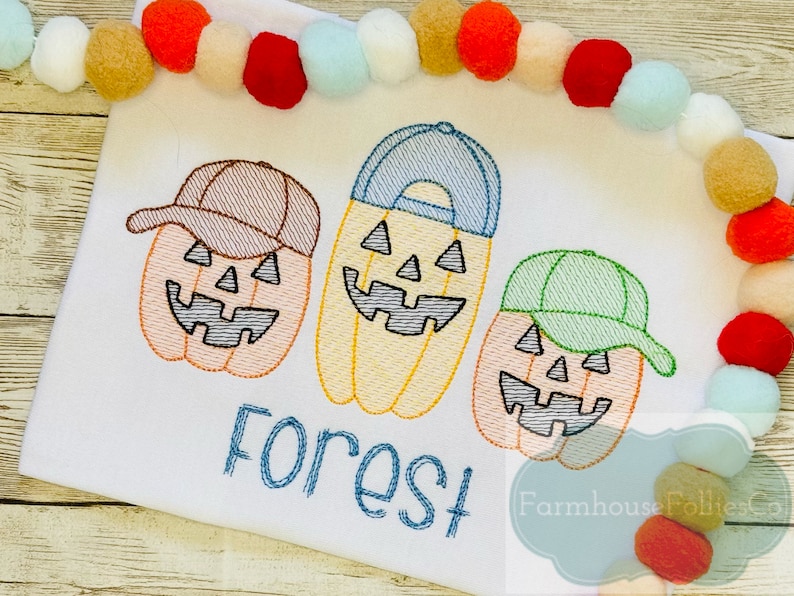 May include: A white t-shirt with three embroidered pumpkin faces wearing baseball caps. The pumpkins are orange, yellow, and peach. The text "Forest" is embroidered below the pumpkins. The text "Farmhouse Follies Co." is embroidered in the bottom right corner.