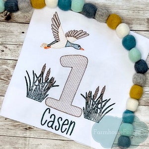 Puede incluir: Un pelele blanco con un dise&ntilde;o bordado de un pato volando sobre un n&uacute;mero uno y juncos. El nombre "Casen" est&aacute; bordado debajo del n&uacute;mero.