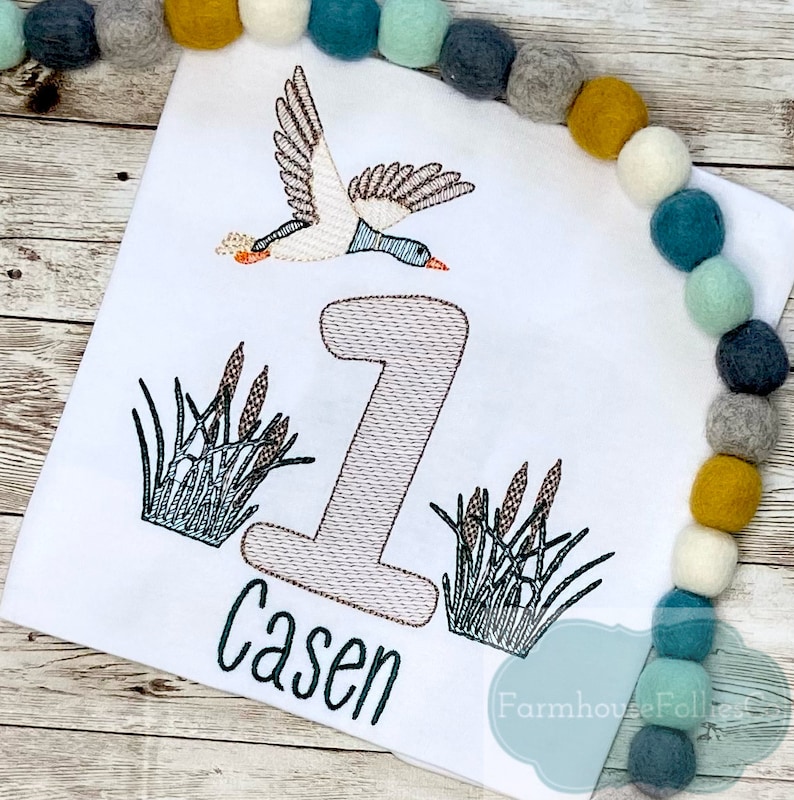 Puede incluir: Un pelele blanco con un dise&ntilde;o bordado de un pato volando sobre un n&uacute;mero uno y juncos. El nombre "Casen" est&aacute; bordado debajo del n&uacute;mero.