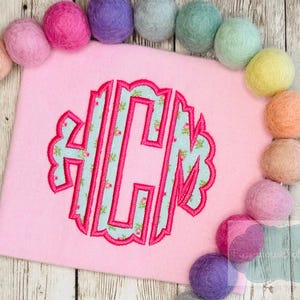 May include: A pink felt square with a floral monogram in the center. The monogram is outlined in a darker pink and filled with a light blue fabric with a floral pattern. Colorful felt balls surround the square.