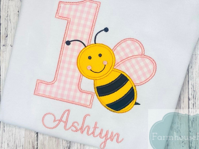 May include: A pink and white gingham number one with a yellow and black bee with pink gingham wings. The name Ashtyn is embroidered below the bee.