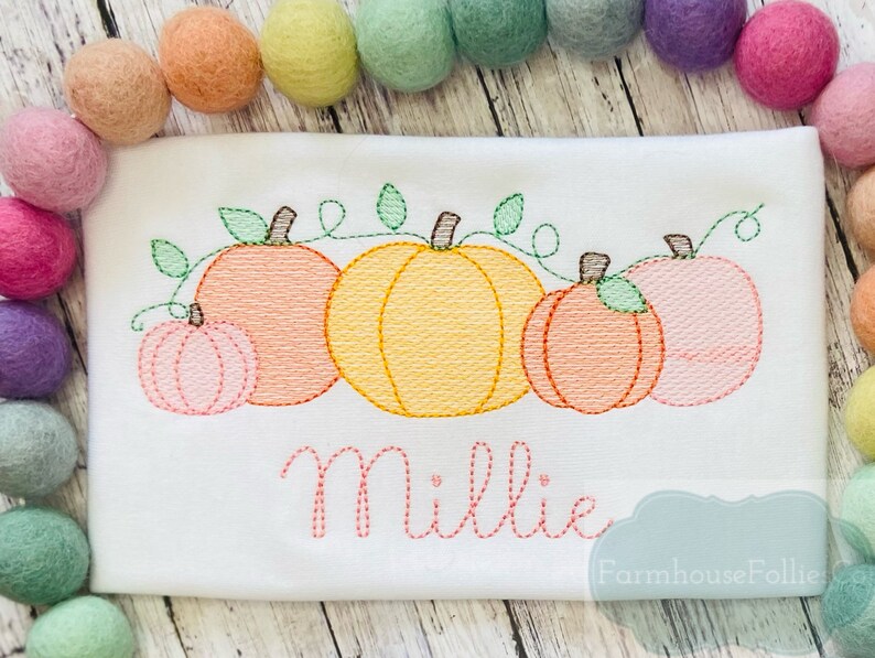 May include: A white onesie with an embroidered design of five pumpkins in a row with green vines. The pumpkins are orange, yellow, and pink. The name "Millie" is embroidered below the pumpkins in pink thread. The text "Farmhouse Follies" is embroidered in the bottom right corner.