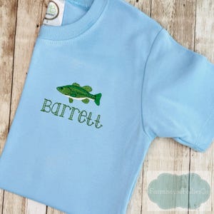 May include: A light blue t-shirt with a green embroidered fish and the name "Barrett" in green thread. The shirt is made of soft material and has short sleeves. A tag is visible near the neckline.