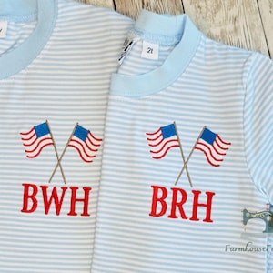 K&ouml;nnte beinhalten: Zwei hellblau-wei&szlig; gestreifte Hemden mit gekreuzten amerikanischen Flaggen auf der Vorderseite. Die Flaggen haben rot-wei&szlig;e Streifen und blaue Felder mit wei&szlig;en Sternen. Die Hemden haben die Buchstaben "BWH" und "BRH" rot gestickt.