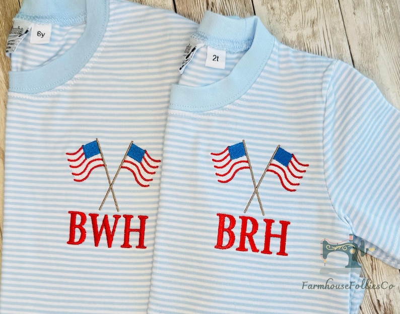 K&ouml;nnte beinhalten: Zwei hellblau-wei&szlig; gestreifte Hemden mit gekreuzten amerikanischen Flaggen auf der Vorderseite. Die Flaggen haben rot-wei&szlig;e Streifen und blaue Felder mit wei&szlig;en Sternen. Die Hemden haben die Buchstaben "BWH" und "BRH" rot gestickt.