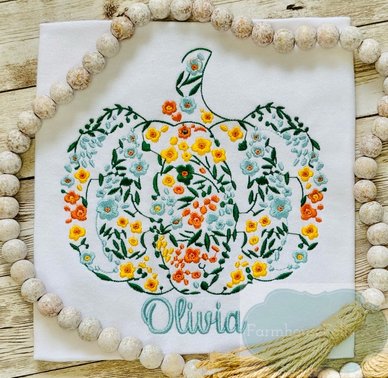 May include: A white t-shirt featuring an embroidered floral pumpkin design in shades of green, yellow, orange, and blue. The name "Olivia" is embroidered below the pumpkin in a light blue thread.