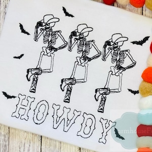 May include: White t-shirt with three black skeleton cowboys wearing hats and boots. The skeletons are in a line and are all facing the right. The word "HOWDY" is written in black below the skeletons. There are black bats flying around the skeletons.