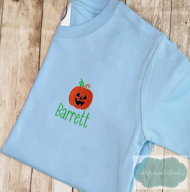 May include: Light blue t-shirt with an embroidered orange pumpkin and the name "Barrett" in green. The pumpkin has a carved face and a green stem. The shirt is made of a soft material.