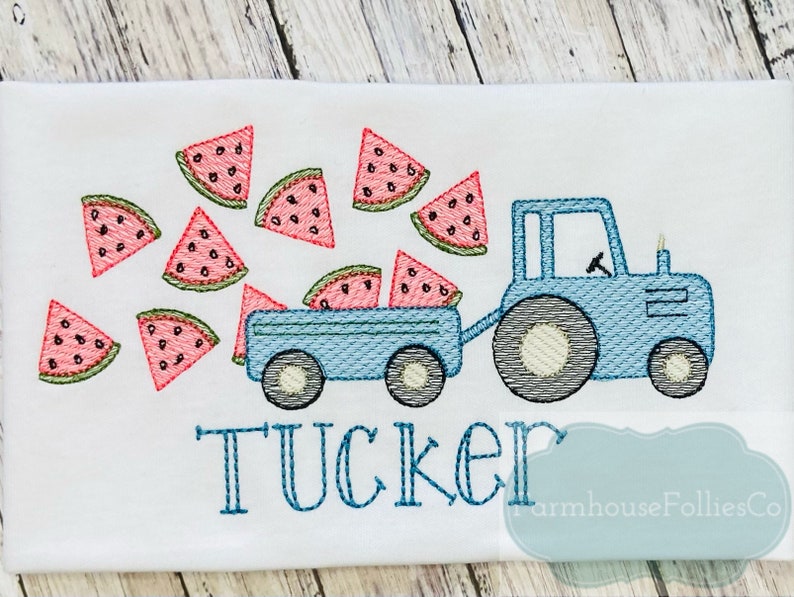 May include: A blue tractor with a wagon full of watermelon slices. The tractor is pulling a wagon full of watermelon slices. The word "TUCKER" is embroidered below the tractor.