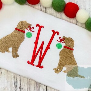 K&ouml;nnte beinhalten: Gestickte Darstellung von zwei braunen Hunden, die rote und gr&uuml;ne Weihnachtskugeln halten, mit einem roten Buchstaben W in der Mitte. Das Design ist auf einem wei&szlig;en Stoffhintergrund.