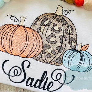 May include: A white t-shirt with an embroidered design of three pumpkins. The pumpkins are orange, blue, and brown with a leopard print pattern. The name "Sadie" is embroidered in a cursive font below the pumpkins.