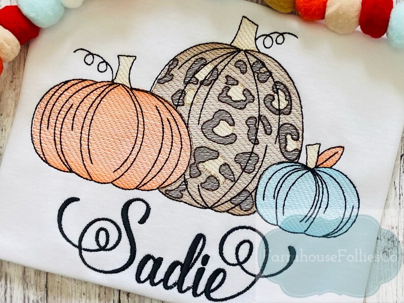 May include: A white t-shirt with an embroidered design of three pumpkins. The pumpkins are orange, blue, and brown with a leopard print pattern. The name "Sadie" is embroidered in a cursive font below the pumpkins.
