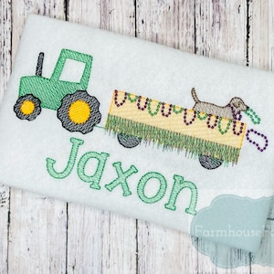 Puede incluir: Tractor verde tirando de un vag&oacute;n con un perro que lleva cuentas moradas y doradas. El vag&oacute;n est&aacute; decorado con hierba verde y cuentas amarillas. El nombre Jaxon est&aacute; bordado en verde debajo del tractor.
