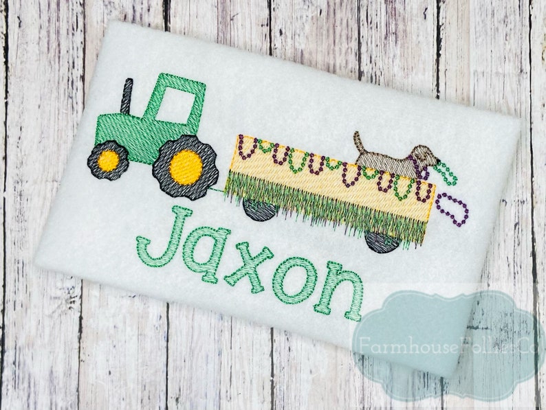 Puede incluir: Tractor verde tirando de un vag&oacute;n con un perro que lleva cuentas moradas y doradas. El vag&oacute;n est&aacute; decorado con hierba verde y cuentas amarillas. El nombre Jaxon est&aacute; bordado en verde debajo del tractor.
